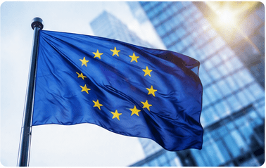 European Union flag waving on a flagpole beside a glass office building with sun flare in the background.